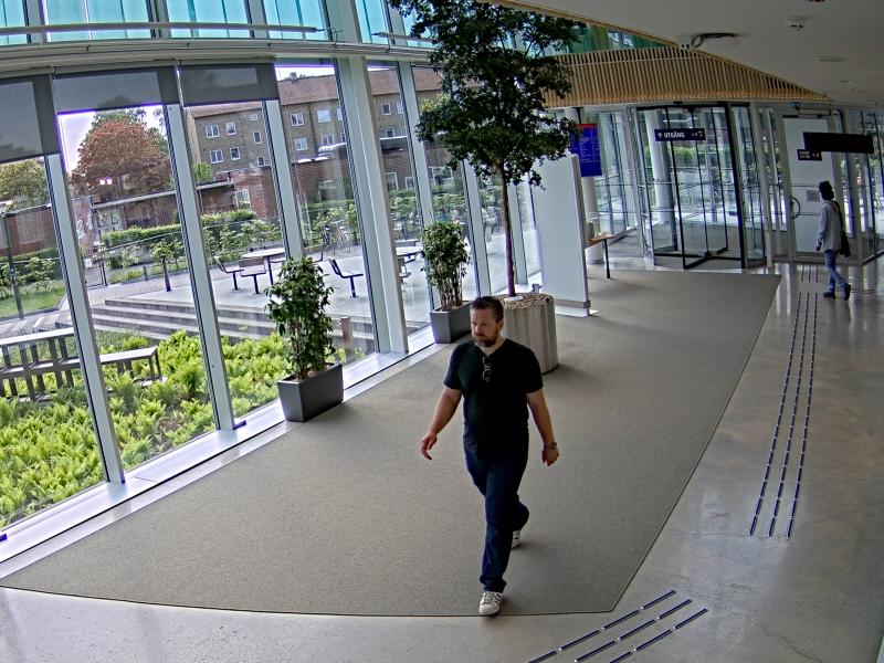 Man seen through a camera lens, walking in a glass corridor