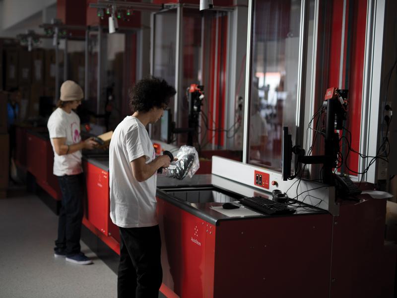 Two men at red warehouse station scanning products, viewed from their right angle.