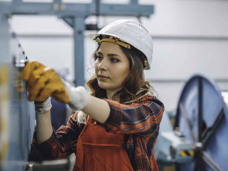 Woman, working in an industrial manufacturing, wearing a white hardhat and red pants