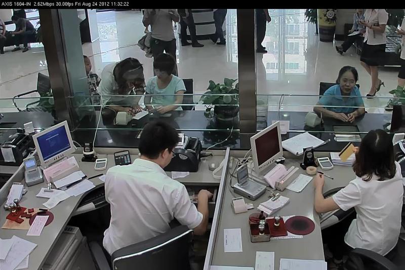 Two people at bank behind glass window assisting people.