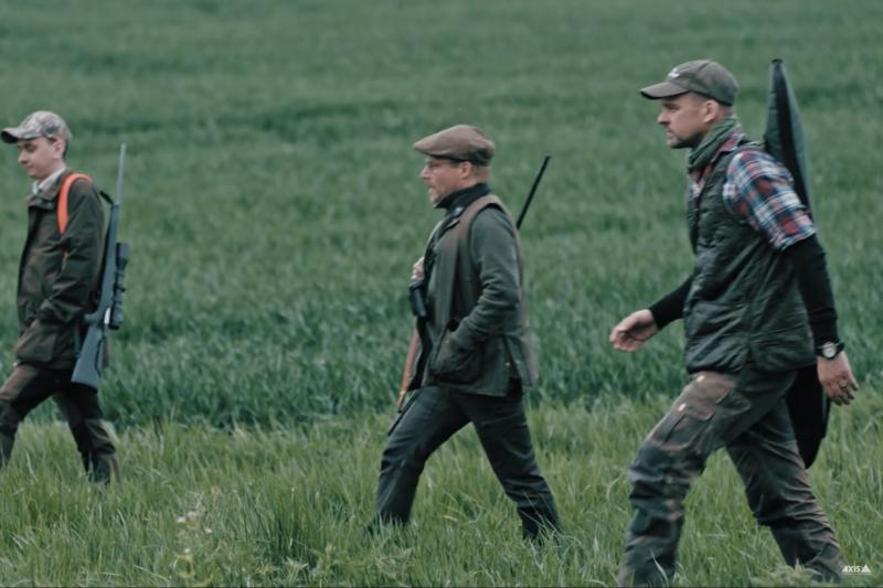 Three people dressed in green walking on a field equipped with hunting gear