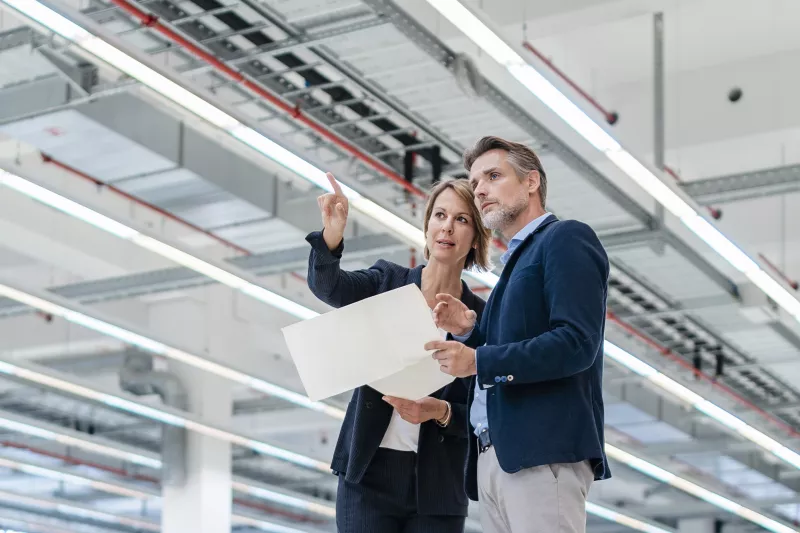 A woman and a man holding a paper and looking over a big space inside a industrial building.