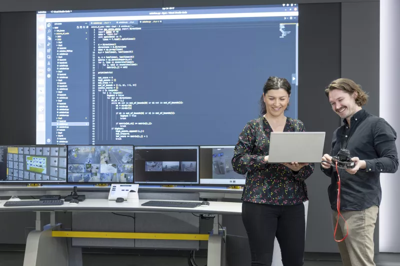 Software engineers looking at a laptop screen. In the background, a big screen with code is showing.