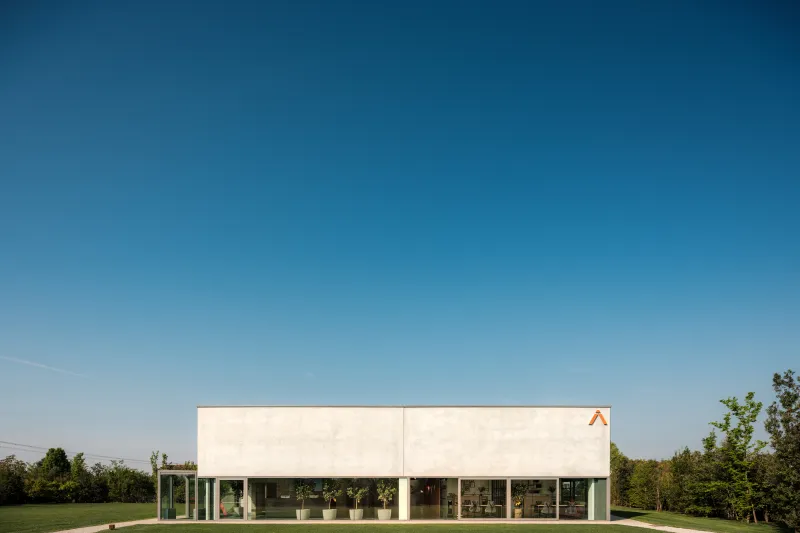 Modern building with a minimalist design, large glass windows, and a white facade, set on a grassy area with a clear blue sky and trees in the background.