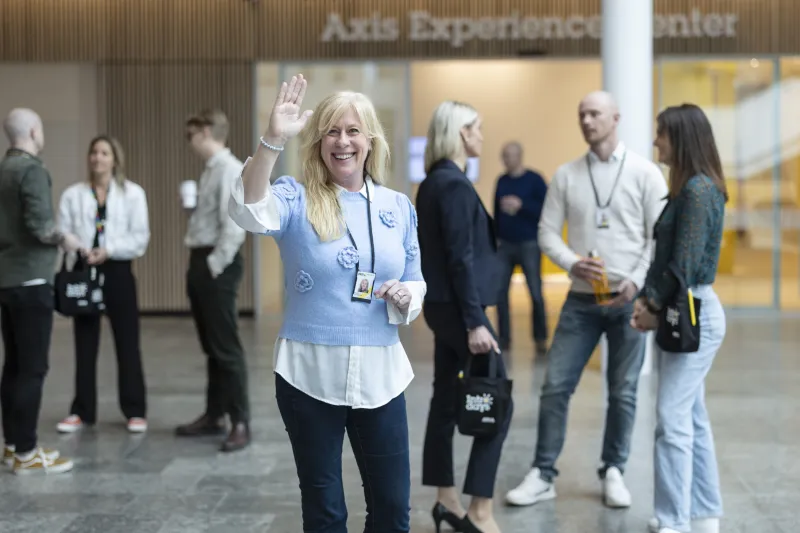 Malin Svensson standing in front of the camera, smiling and waving, with people standing in the background talking.