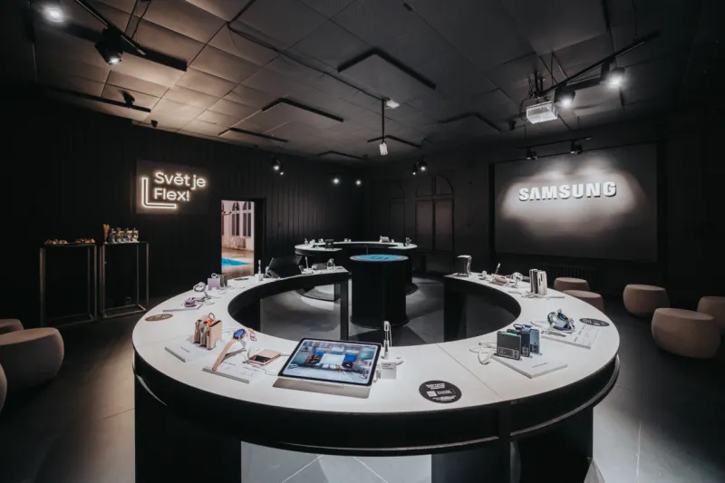 A sleek, modern Samsung showroom featuring minimalist white circular display tables with various gadgets, set against a moody, dark background with spotlights.