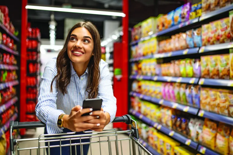 A happy woman, smartphone in hand, walking around a grocery store with a shopping cart. 