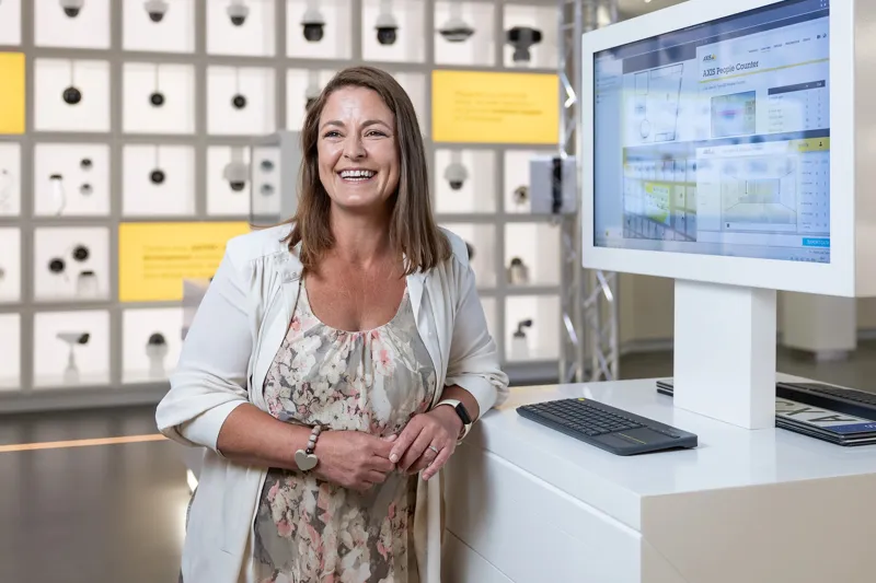 Vanessa Tyne, Strategic Customers Technical Manager at Axis, standing infront of the product wall in Axis Experience Center, smiling. 