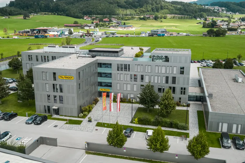 An aerial view of a modern gray multi-story Palfinger office building surrounded by a parking lot, green lawns, and a scenic, rolling rural landscape.