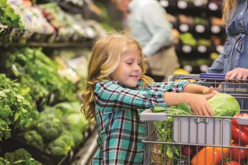 A child putting lettuce in their shopping cart amongst other vegetables.
