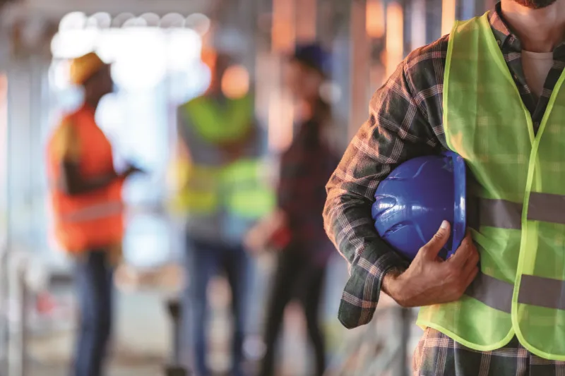 A construction worker holding a blue protection helmet with blurry scenery behind him.