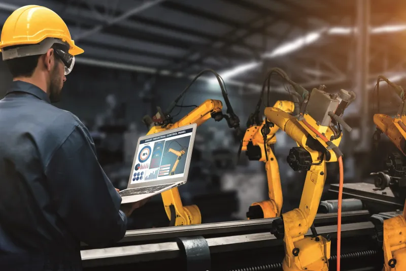 Man with a hardhat on, standing in front of a laptop and robots.