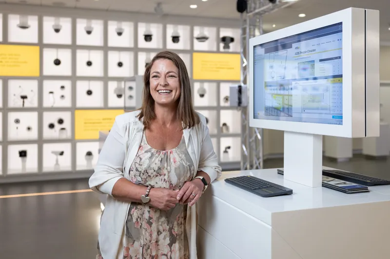 Vanessa Tyne, Strategic Customers Technical Manager at Axis, standing infront of the product wall in Axis Experience Center, smiling. 