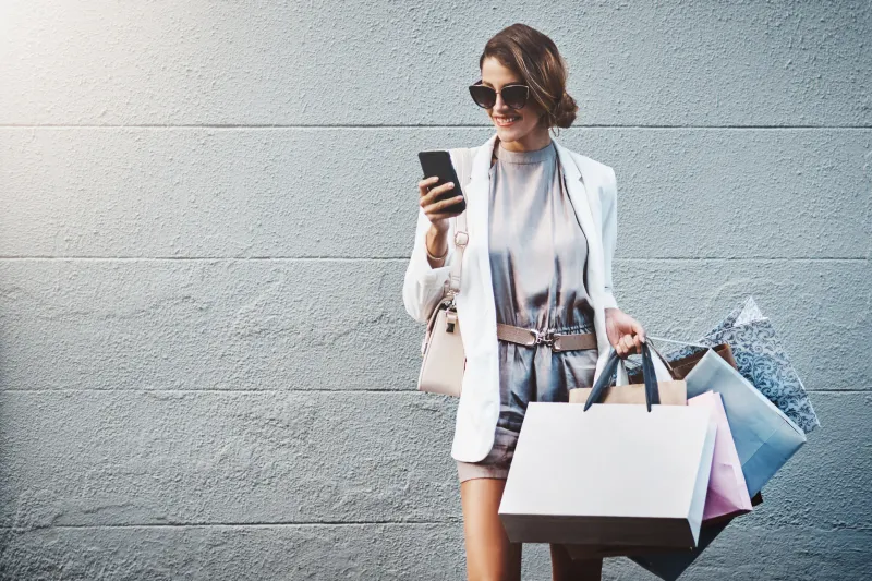 A woman holding her smartphone and smiling at the screen. She has sunglasses on and multiple shopping bags.