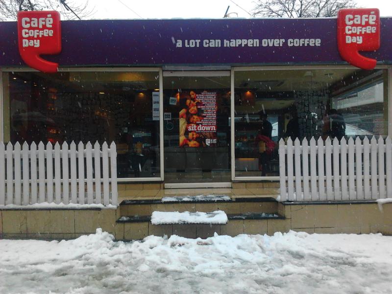 Outside of Cafe coffe day, purple and red signs and white fence in front.