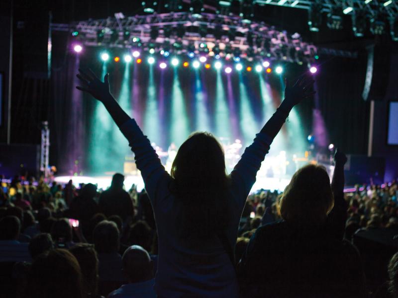 Woman with raised arms at concert, only light is on stage.