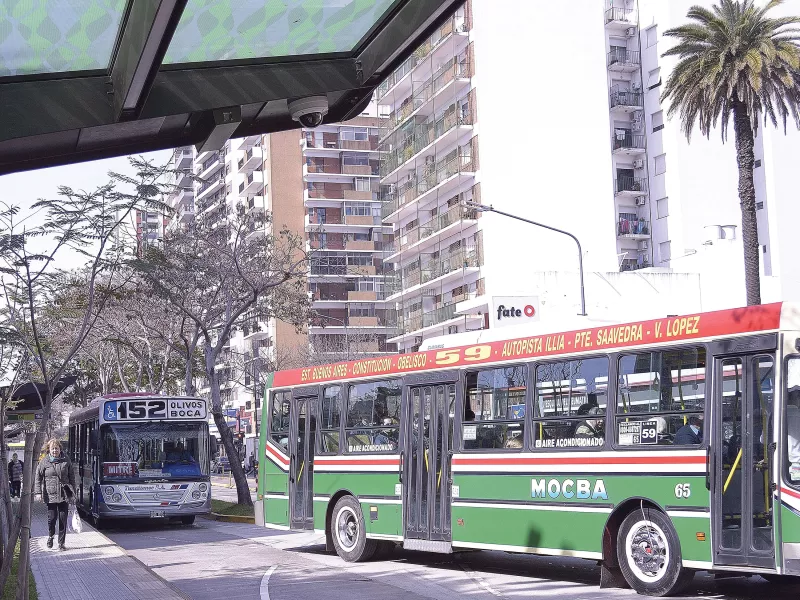 Bus stop with one green and one blue bus, apartment building in background.