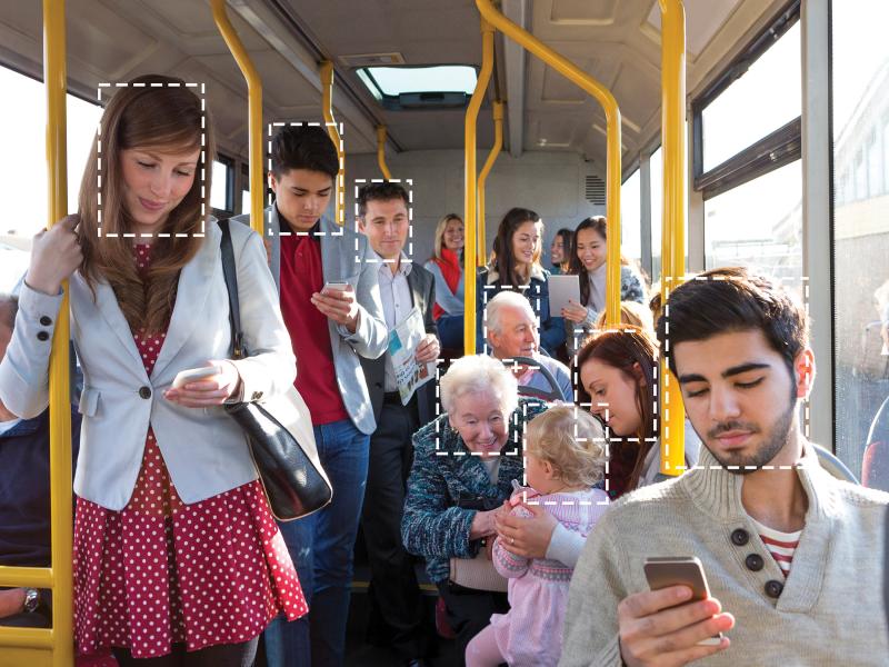 Crowded on a bus, several of the passangers are looking at their phone. Peoples face are marked