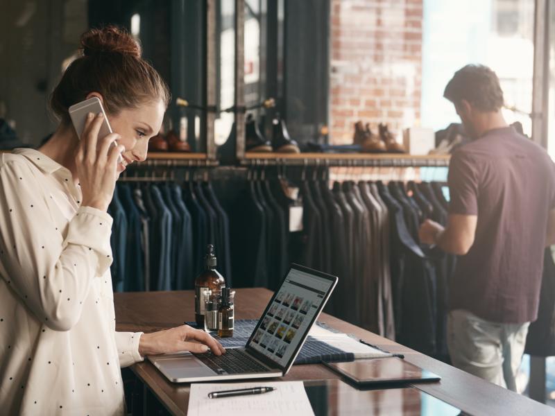 Women behind desktop in a clothes store making a call and looking at her laptop