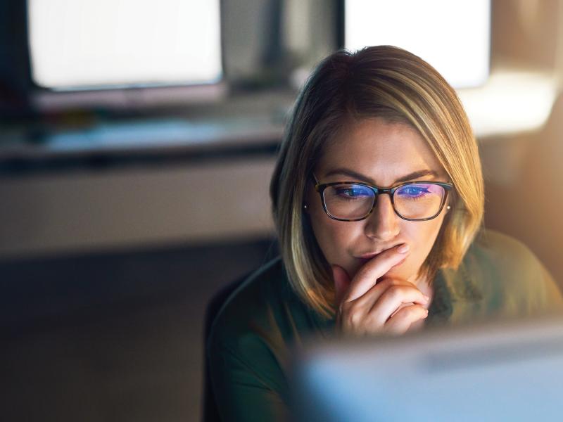 Woman wearing glasses, sitting by a computer thinking