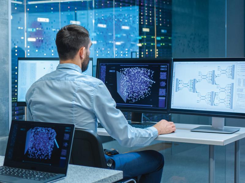 a man who sitting at his desk, monitoring three computer screens