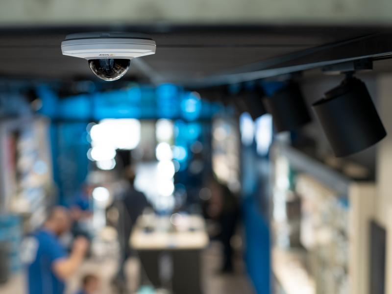 Черно-белая камера M5075 с логотипом AXIS висит на потолке в розничном магазине.