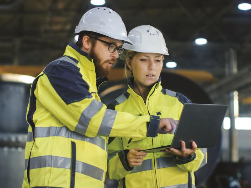 man and woman who are wearing hardhats and safety vests, looking at a tablet together