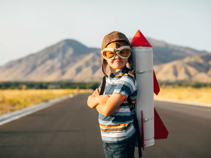boy with a rocket on his back, standing on a road. mountains in the background.