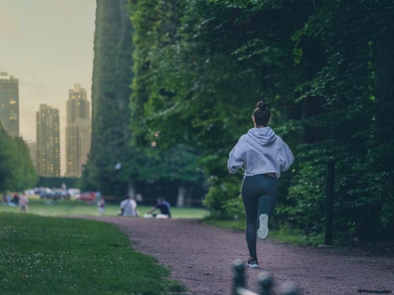 Woman running in a park. Foliage to the right and high-rise buildings to the left.