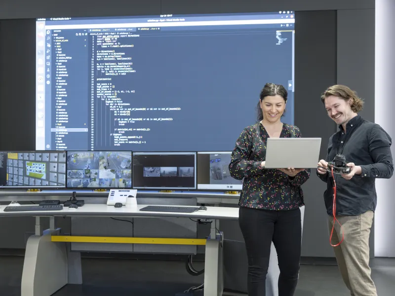 Software engineers looking at a laptop screen. In the background, a big screen with code is showing.