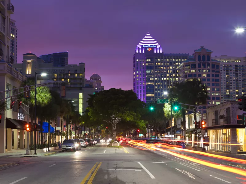 Overview of city street during early nighttime, the sky is a deep purple and the lights from the skyline sparkles 