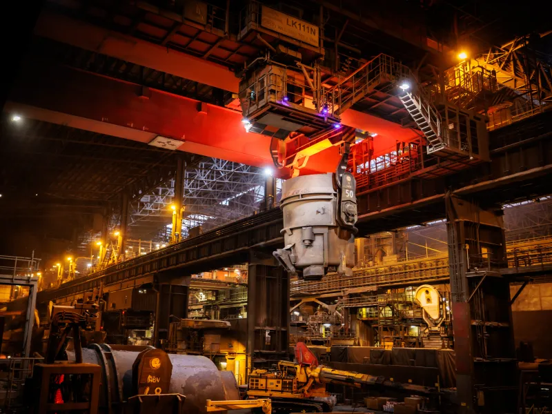 Overview of a ladle with molten steel suspended from ArcelorMittal's manufacturing facility's roof  lifted by cranes. 