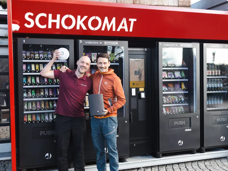 Two people smiling by a red "SCHOKOMAT" vending machine; one holds a white IP speaker, the other a black network speaker