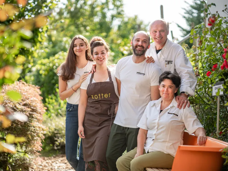 Five people in a lush garden, with one woman wearing a Zotter apron, surrounded by greenery and an orange container.