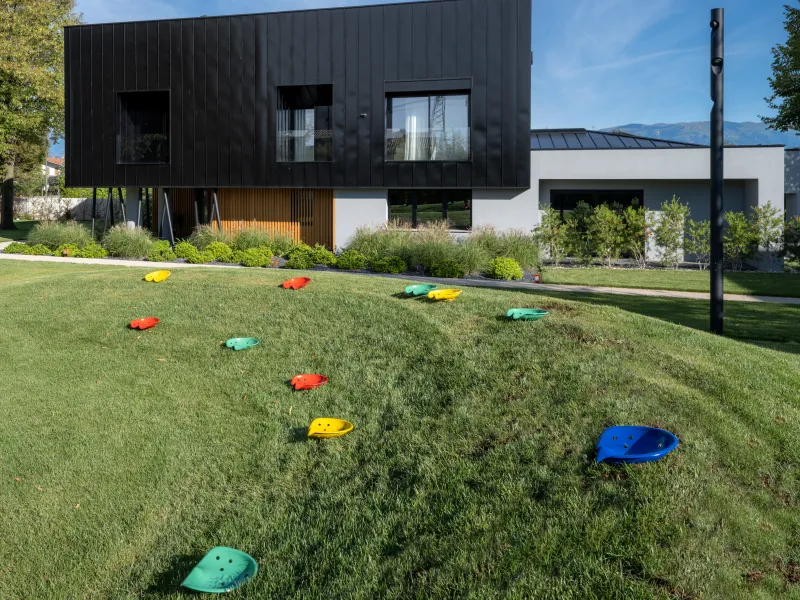  Modern black two-story building on stilts with colorful stepping stones on a grassy slope.