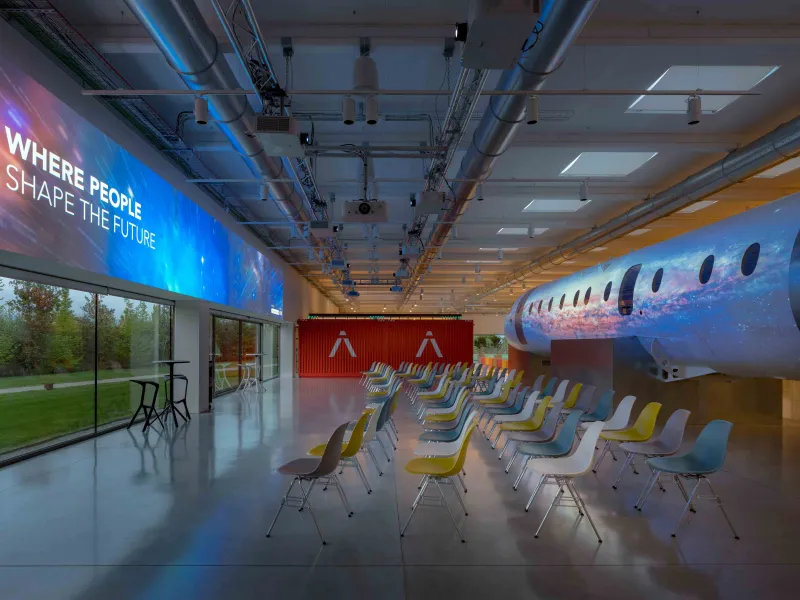 A modern conference room with a futuristic design, featuring a large digital screen displaying "WHERE PEOPLE SHAPE THE FUTURE," an airplane fuselage-like structure with space images, colorful chairs, and large windows with a view of greenery.