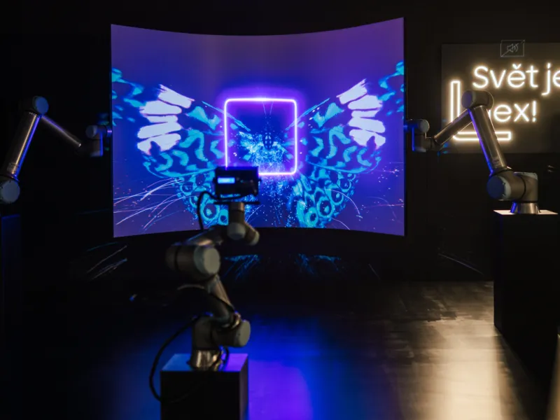 A futuristic art installation featuring robotic arms, a vibrant curved screen, and a glowing "Svět je lex!" sign