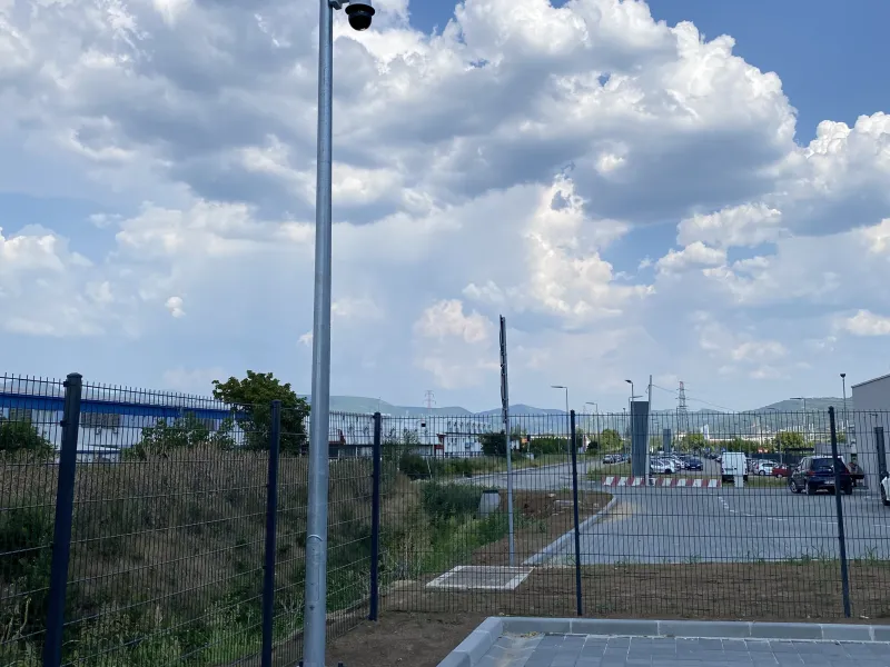 A security camera mounted on a tall metal pole stands near a fenced perimeter, overlooking a parking area with a distant road under a cloudy sky.
