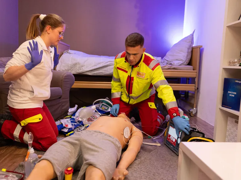 Two emergency responders practicing medical protocols on a training mannequin in a simulated room.