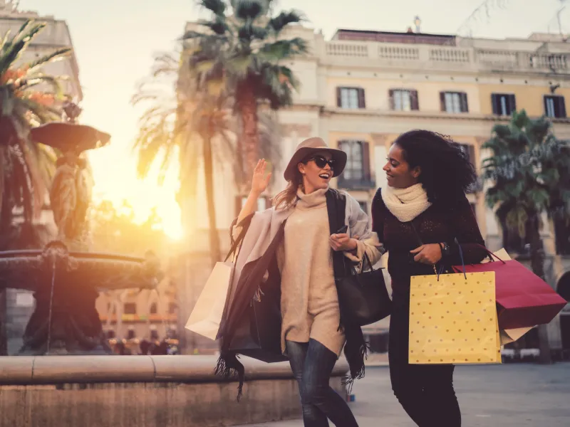 Two women shopping outdoors and walking past a fountain.
