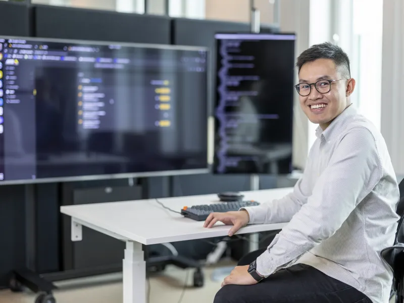 Axis employee Andy Truong smiling, sitting by his desk in front of two large screens.