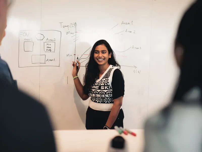 An Axis student worker holding a pen and standing in front of a whiteboard, giving a presentation to other Axis colleagues.