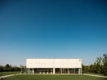  Modern building with a minimalist design, large glass windows, and a white facade, set on a grassy area with a clear blue sky and trees in the background.