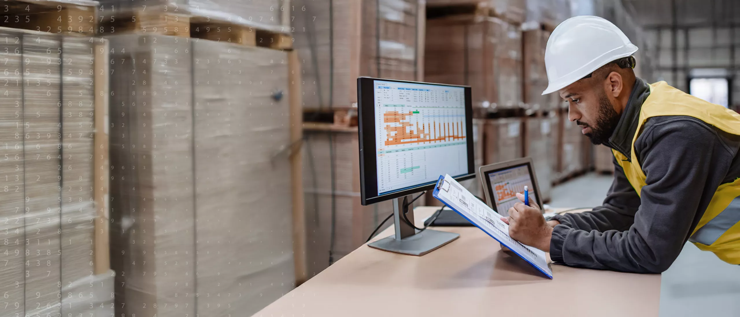 A construction worker holding a clipboard over a desk with a computer.