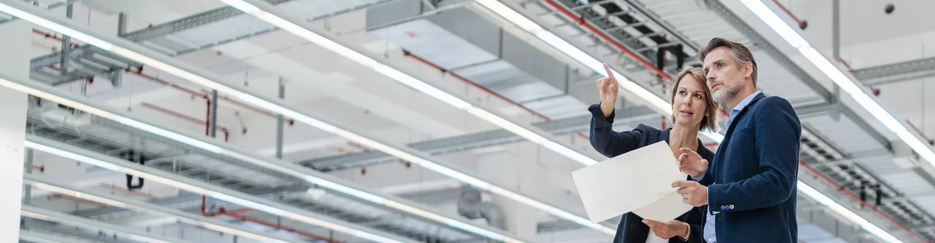 A woman and a man holding a paper and looking over a big space inside a industrial building.
