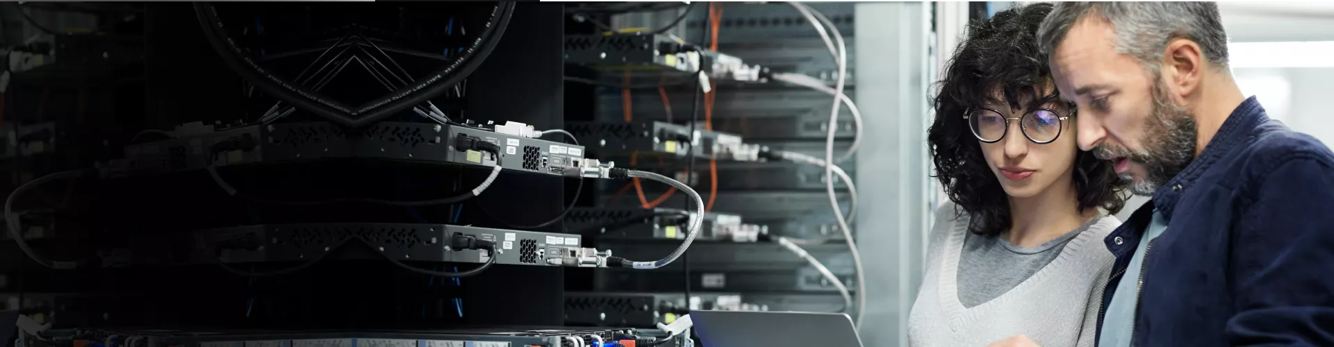 woman and man standing next to server racks discussing, with laptop and tablet in their hands