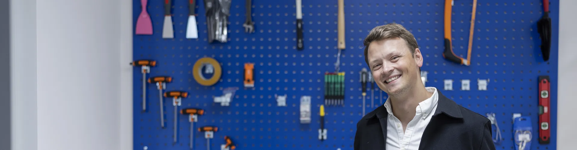 A man standing and smiling in front of a wall with tools hanging from it.