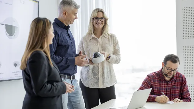 Four people in a meeting room having a conversation about an Axis camera. There's a laptop on the table and a screen behind them.