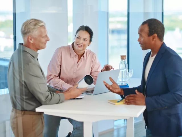 Two men, one holding an Axis camera, and a woman standing around the table having a conversation in the office.