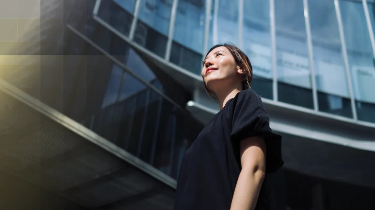 Asian woman looking up at the sky with a smile. A close up of a glass building behind her.